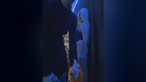 Media: Video of a young woman with long, straight white hair, wearing a white dress, standing in a dimly lit, dark room with a glowing blue light source in the background.