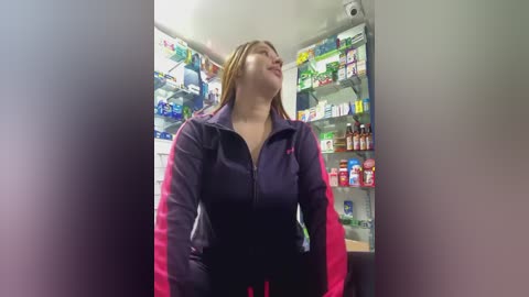 Media: Video of a smiling Asian woman with shoulder-length hair, wearing a black and pink jacket, standing in a brightly lit pharmacy with shelves full of medicine bottles.