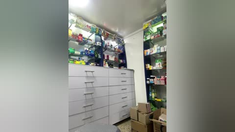 Media: Video of a small, cluttered room with a white cabinet, multiple shelves filled with colorful boxes, and scattered cardboard boxes on the floor.