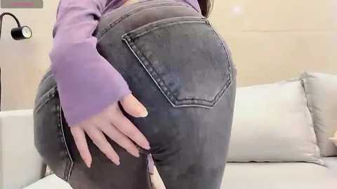 Media: Video of a woman with fair skin, wearing a purple long-sleeve top and tight, dark blue jeans. She is bent over a white couch, her hand resting on her rear.