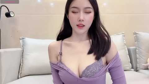 Media: Video of an East Asian woman with long black hair, fair skin, and a slender physique, wearing a purple off-shoulder top exposing a lavender lace bra, sitting on a white bed with light blue pillows in a modern, softly lit bedroom.