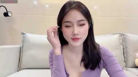 Media: Video of a young East Asian woman with straight black hair, fair skin, and light purple top, adjusting her earbud, seated on a beige couch with decorative pillows.