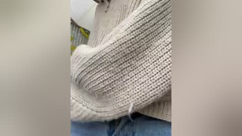 Media: Video of a beige, textured, knitted sweater with a slightly torn edge, partially covering a person's upper body. The background features a blurred, light-colored surface with indistinct objects, possibly furniture or fabric.