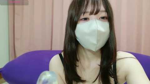 Media: Video of a young Asian woman with long black hair and fair skin, wearing a white face mask, black tank top, and sitting on a purple pillow.