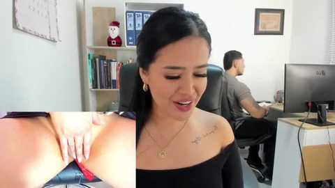 Media: Video of a woman with medium skin tone and black hair in an office, wearing a black off-shoulder top, exposing her red lace bra. A man works at a desk behind her.