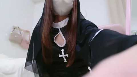 Media: A video of a woman in a black nun's habit with a white cross, wearing a surgical mask, sitting on a bed with a beige bag in the background.