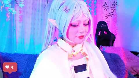 Media: Video of a young Asian woman in a white cosplay outfit, with light blue hair and elf ears, surrounded by vibrant blue and pink lights, holding a black microphone.