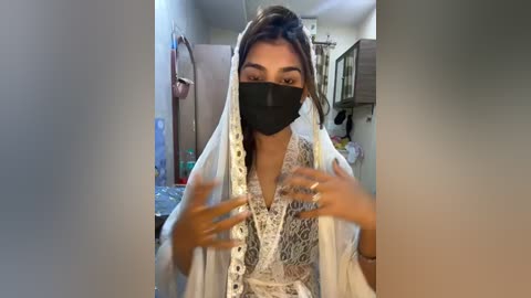 Media: Video of a South Asian woman with medium skin tone, wearing a white veil with lace detailing, a black mask, and a floral-patterned blouse. She is indoors, in a cluttered room with kitchen items and a hanging pot in the background.