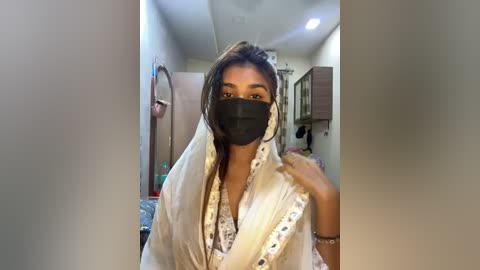 Media: Video of a South Asian woman with medium-brown skin, wearing a white dupatta with blue and white patterns, black mask, and black face shield, standing indoors in a cluttered room with a beige wall and a white ceiling fan.