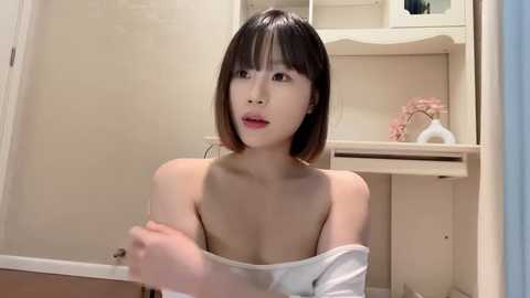 Media: Video of an Asian woman with straight, shoulder-length black hair, fair skin, and a slender physique. She is topless, wearing a white off-shoulder top, in a cream-colored bathroom with a white sink and pink flowers.