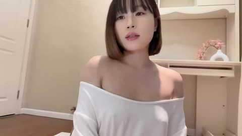 Media: A video of an East Asian woman with a bob haircut, wearing a white off-shoulder top, sitting on the floor in a minimalist, beige-walled room with a desk and pink decorative item in the background.