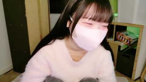 Media: Video of an Asian woman with long black hair, wearing a white face mask, a white sweater, and a grey blanket, sitting on a beige carpeted floor, indoors with a wooden cabinet and green-lit background.