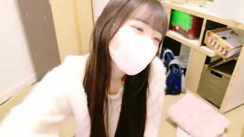 Media: Video of an Asian woman with long black hair, wearing a white mask, and a white fur-lined coat, kneeling in a cluttered, beige room with shelves and baskets.