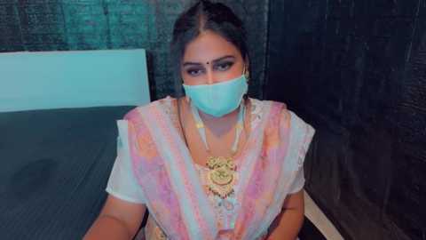 Media: Video of a woman with a fair complexion, dark hair, and a light blue mask, wearing a colorful saree with a golden pendant, seated in a dimly lit room with dark brick walls.