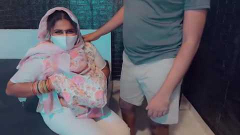 Media: Video: A woman with a mask and pink headscarf, wearing a floral blouse and gold bangles, sits on a dark bench. A man in a gray shirt and white shorts stands next to her, holding her arm.