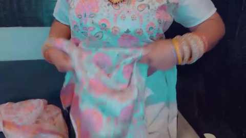 Media: Video of a woman in a teal and pink floral dress with a white underskirt, holding a pink shawl. She wears gold bangles and a necklace, standing indoors against a dark background.