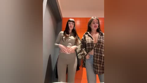 Media: Video of two women, one in a beige jumpsuit, the other in a plaid shirt, standing in a narrow corridor with orange walls and a mirrored door.