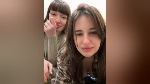 Media: Video of a young woman with long, wavy brown hair and a serene expression, wearing a patterned jacket. She is seated beside a younger girl with straight, dark hair, who is touching her face with a contemplative expression.