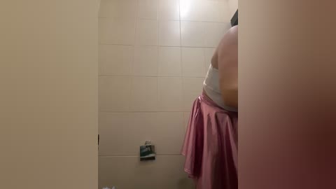 Media: Video of a person in a shower stall, wearing a pink towel draped over their shoulder, with light beige tiles and a metallic showerhead visible.