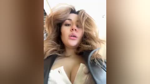 Media: Video of a young woman with voluminous, curly blonde hair, light skin, and full lips, wearing a black leather jacket, partially revealing a white blouse.