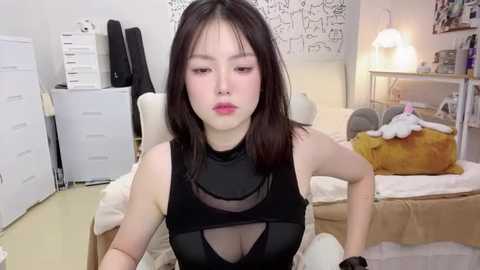 Media: Video of an Asian woman with straight, shoulder-length black hair, wearing a sheer black top, sitting on a beige chair in a cluttered, minimalistic bedroom.