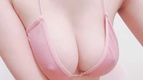 Media: Close-up video of a fair-skinned woman's chest in a light pink, V-neck bra, revealing ample cleavage. The background is plain white, emphasizing the bra's delicate texture and the woman's smooth skin.