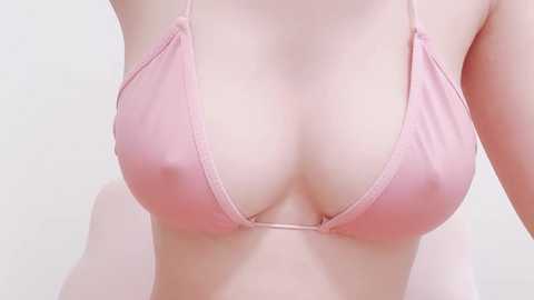 Media: Video of a woman's bare chest in a pale pink triangle bikini top, revealing moderate cleavage and a smooth, light-skinned complexion. The background is a plain, off-white wall.