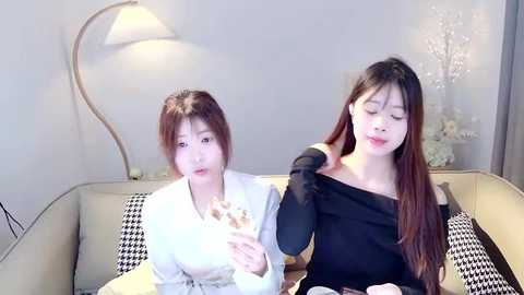 Media: Video of two young Asian women, one in a white shirt and another in a black off-shoulder top, sitting on a beige sofa, eating ice cream, with a modern lamp in the background.