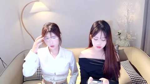 Media: Video of two Asian women in a modern living room. One wears a white shirt, the other a black off-shoulder top. They sit on a beige sofa with houndstooth pillows, under a curved floor lamp.