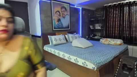 Media: Video of a modern bedroom with a woman in a green blouse, a couple in a framed photo, a bed with blue sheets, and dark curtains.