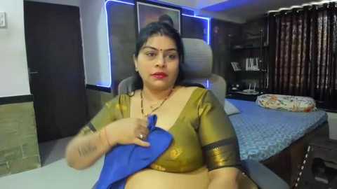 Media: Video of a plus-sized South Asian woman with dark hair and medium skin tone, wearing a gold blouse and blue sari, seated at a desk in a modern office with blue lighting, bookshelves, and a bed.