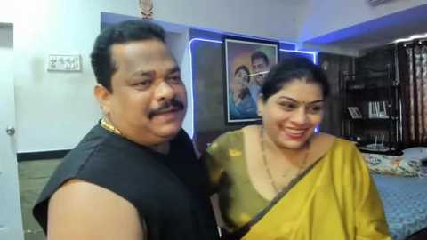 Media: A video shows a middle-aged South Asian couple smiling in a modern living room with framed portraits, bookshelves, and blue LED strip lighting. The man wears a black shirt, and the woman a mustard-yellow sari.