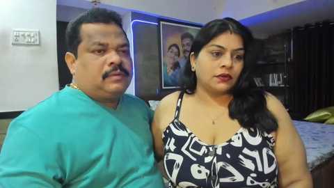 Media: Video of a middle-aged South Asian man and woman in a living room. The man, with short dark hair and a mustache, wears a teal shirt. The woman, with long black hair, a black-and-white patterned dress, and red lipstick, gazes at the camera.