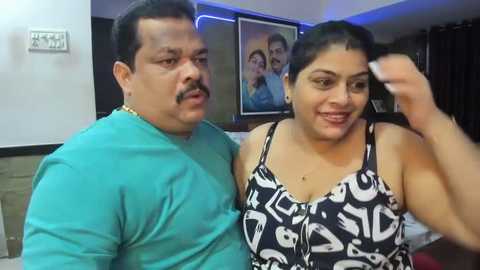 Media: Video of a middle-aged South Asian couple in a living room. The man, with a mustache, wears a teal shirt, and the woman, with dark hair, dons a black top with white abstract designs.