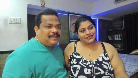 Media: Video of an overweight South Asian man with mustache in a teal shirt and a plus-size woman in a black and white patterned dress, standing indoors.