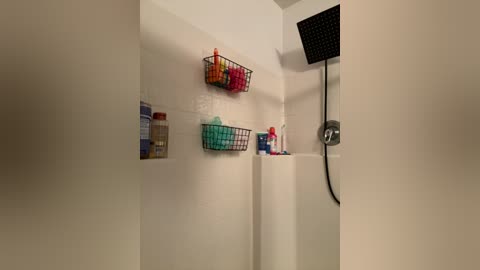 Media: Video of a minimalist, modern shower with white tiles, featuring a black rain showerhead, two wire baskets with toiletries, and a silver showerhead holder.