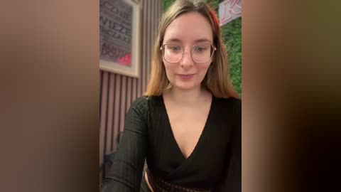 Media: Video of a young Caucasian woman with fair skin and light brown hair, wearing glasses and a black V-neck top, standing in a room with vertical striped walls and a green plant in the background.