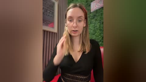 Media: Video of a young Caucasian woman with light skin, straight blonde hair, wearing glasses and a black V-neck top, looking surprised and holding her hand to her face. Background features greenery and a red couch.