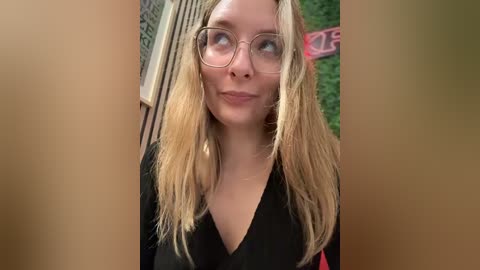 Media: Video of a fair-skinned woman with long, blonde hair, wearing glasses and a black shirt, standing against a green wall and wooden paneling.