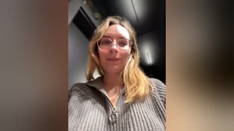 Media: Video of a young Caucasian woman with shoulder-length blonde hair, wearing clear glasses and a gray knit zip-up sweater. She has a slight smile and stands indoors with a blurred background.