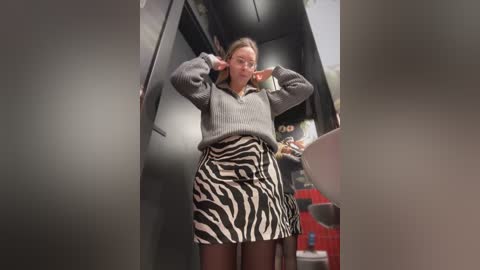 Media: Video of a woman in a gray sweater, black tights, and a zebra-patterned skirt, posing with arms raised, inside a narrow, dimly lit hallway with gray walls and a red object in the background.