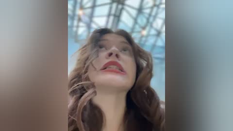 Media: A video of a young woman with fair skin, wavy brown hair, and red lipstick, looking upward. The background features a blurred, modern, glass-paned ceiling.