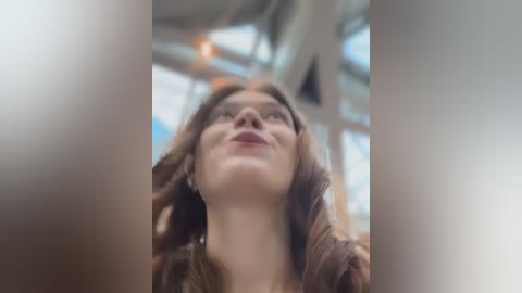 Media: Video of a young woman with wavy, light brown hair, wearing a brown coat, looking upwards with a serene expression, in a modern, sunlit interior with large windows and white beams.