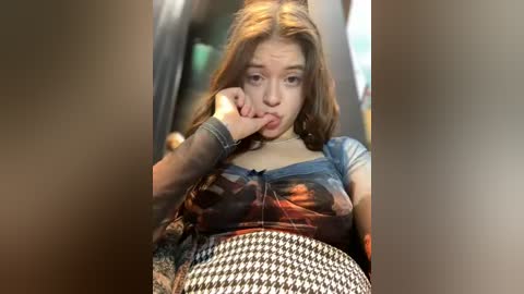 Media: Video of a young woman with fair skin, long brown hair, and a plaid shirt, holding her chin in thought, sitting on a black and white houndstooth-patterned cushion.