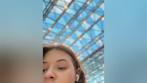 Media: Video of a young woman with light skin and long brown hair, wearing white headphones, gazing upwards at a modern, glass-roofed building with geometric patterns.