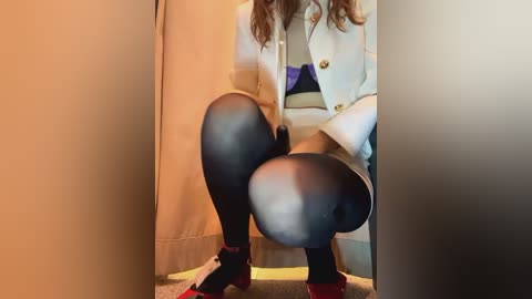 Media: Video of a woman squatting, wearing a white blazer and black leggings, with red shoes, against a beige curtain. Her legs are prominent, with the camera angle emphasizing her thighs and buttocks.