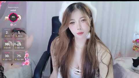 Media: Video of a young East Asian woman with long brown hair, fair skin, and a slender physique, wearing a white dress and a fluffy hat. Background shows a gaming setup with a dark chair and a monitor displaying a virtual avatar.