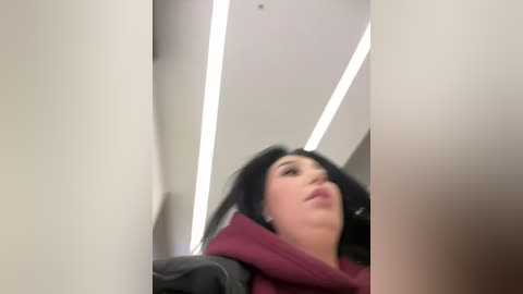 Media: A blurred video of a woman with long black hair, fair skin, and wearing a burgundy hoodie, captured from a low angle in a dimly lit hallway with white walls and fluorescent lights.