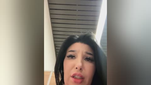 Media: A video of a Latina woman with long, dark hair, dark eyes, and full lips, wearing makeup, captured from a low angle in an indoor hallway with a tiled ceiling.