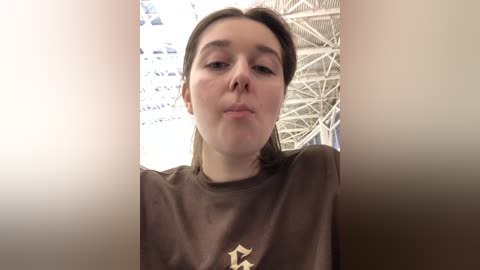 Media: Video of a young woman with light skin and brown hair, wearing a brown t-shirt, blowing a kiss. Background shows a white ceiling with blue patterns.